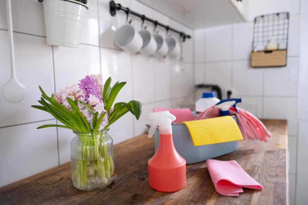 Comptoir de cuisine avec des produits de nettoyage, des gants roses, des chiffons colorés et un bouquet de jacinthes printanières.