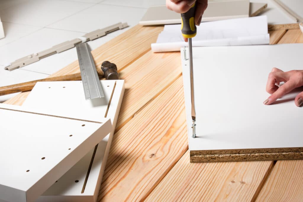 Main vissant une vis dans un meuble en kit blanc, tournevis "CHROME VANADIUM". Outils, instructions et panneaux de bois sur table.