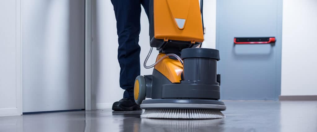 Opérateur nettoyant un couloir avec une lustreuse de sol professionnelle orange et grise, laissant le carrelage brillant.